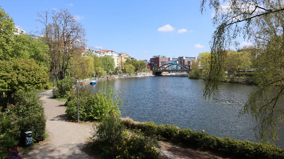 Grünes Flussufer mit Spazierweg, einer Brücke und blauem Frühlingshimmel.